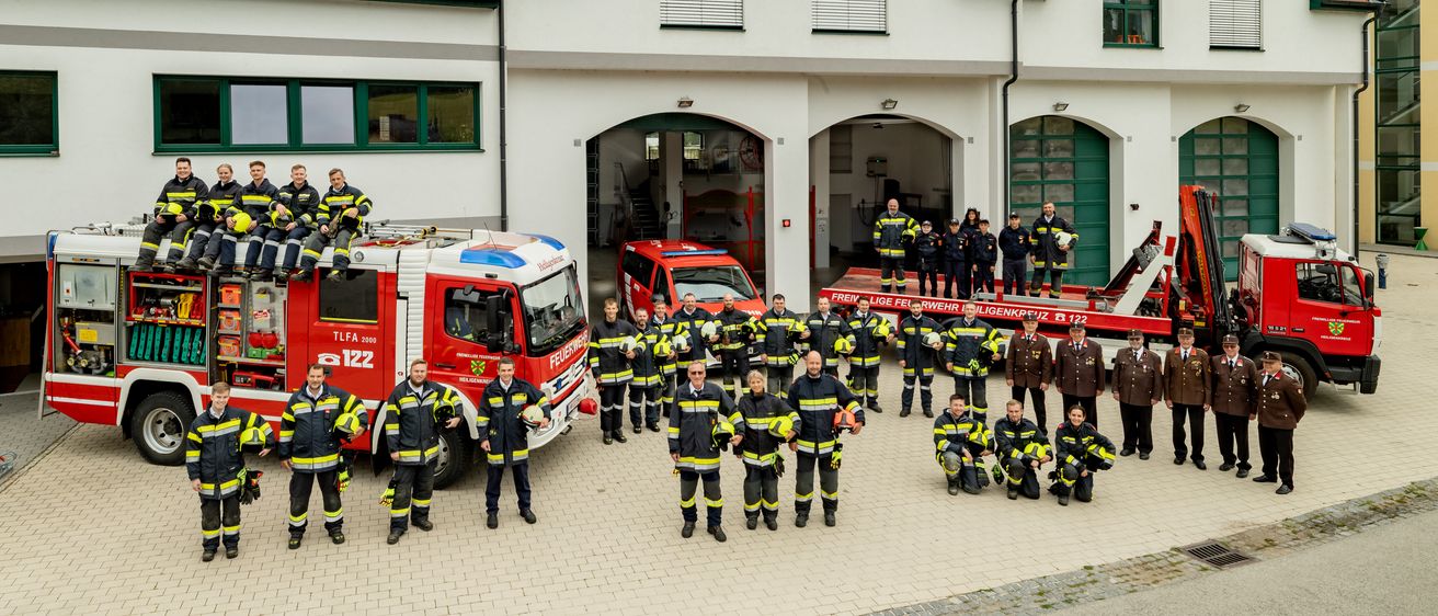Eine Gruppe von Feuerwehrleuten, einige stehend und andere kniend, posiert vor einem Feuerwehrhaus. Sie tragen gelbe und schwarze Uniformen und Helme. Im Hintergrund sind zwei Feuerwehrwagen geparkt.