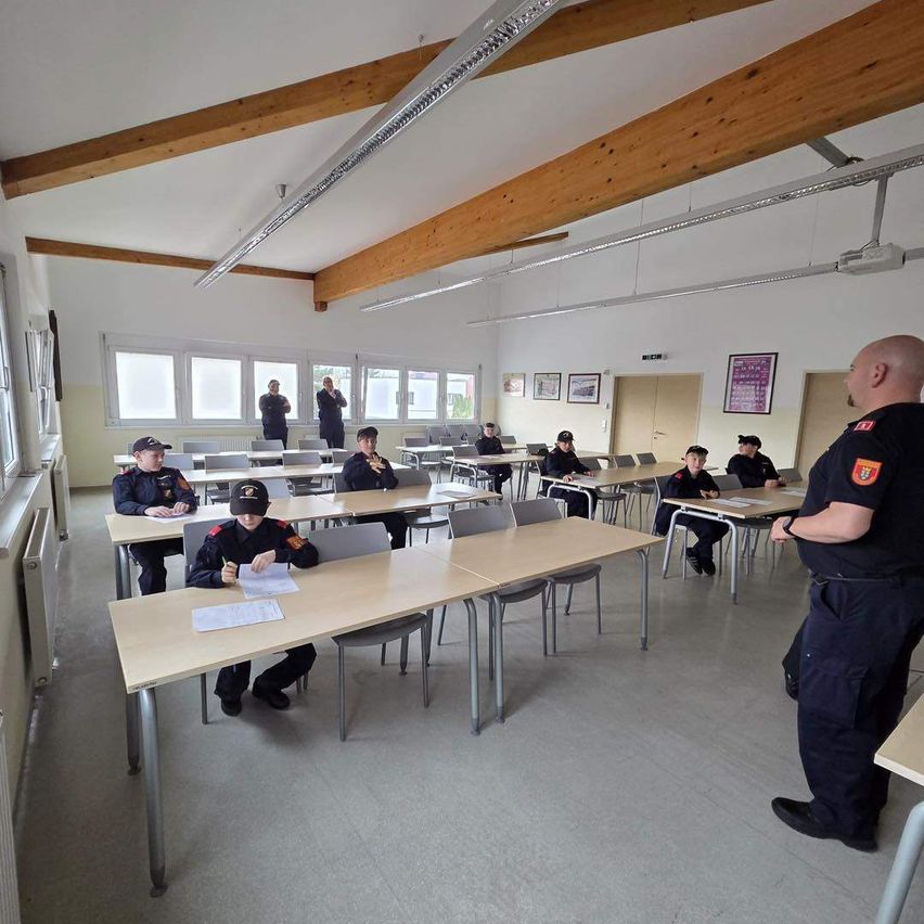 Ein Klassenzimmer mit uniformierten Personen, die sitzen, und einem Polizeibeamten, der Anweisungen gibt. Holzbalken und Fenster schmücken den Raum.