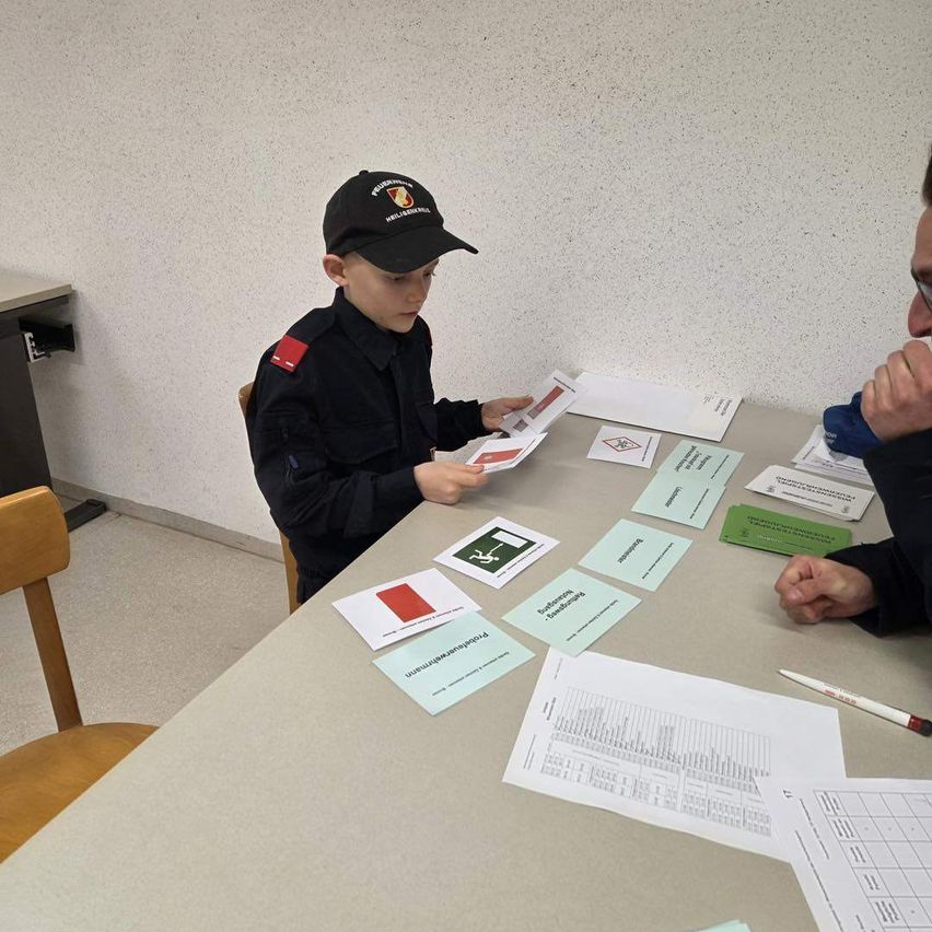 Ein Junge in Uniform hält eine rote und weiße Karte und sitzt an einem Tisch mit mehreren Karten und Papieren. Ein erwachsener Mann sitzt neben ihm. Der Raum hat eine schlichte weiße Wand.