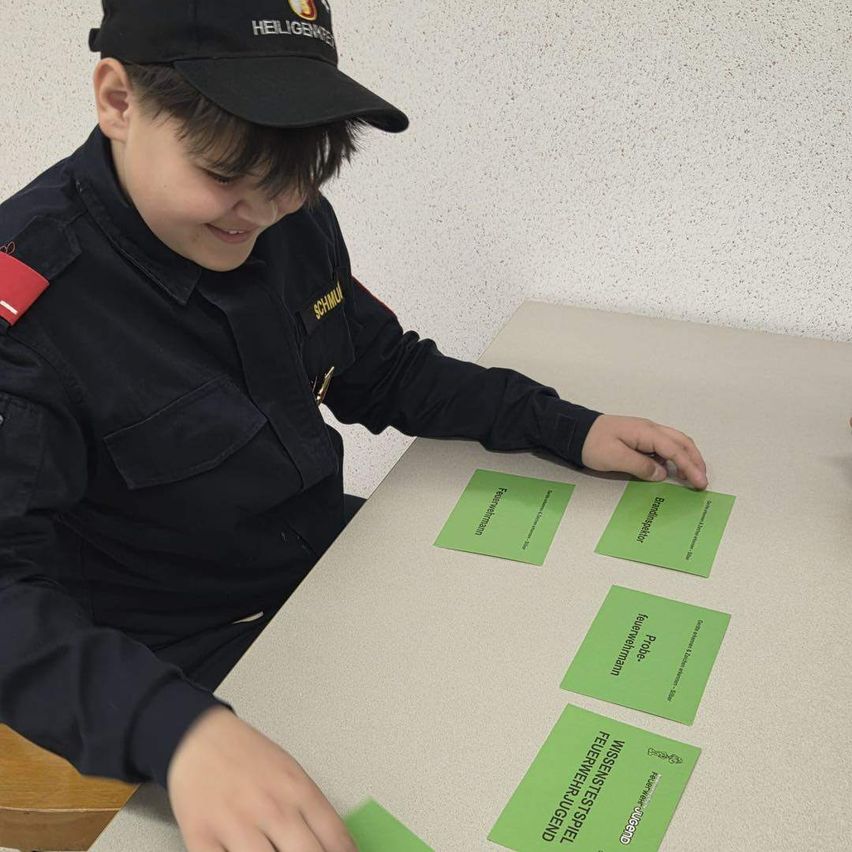 Ein junger Mann in Uniform sitzt an einem Tisch, lächelt und schaut auf grüne Karten mit Worten darauf. Die Karten haben Wörter wie 'Wissenstest' und 'Probenahme'. Die Wand hinter ihm ist weiß.