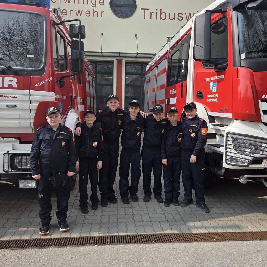 Eine Gruppe junger Leute in Uniformen steht vor zwei roten Feuerwehrwagen. Sie alle lächeln. Dahinter steht ein Gebäude mit dem Wort 'Tribuswi' darauf.