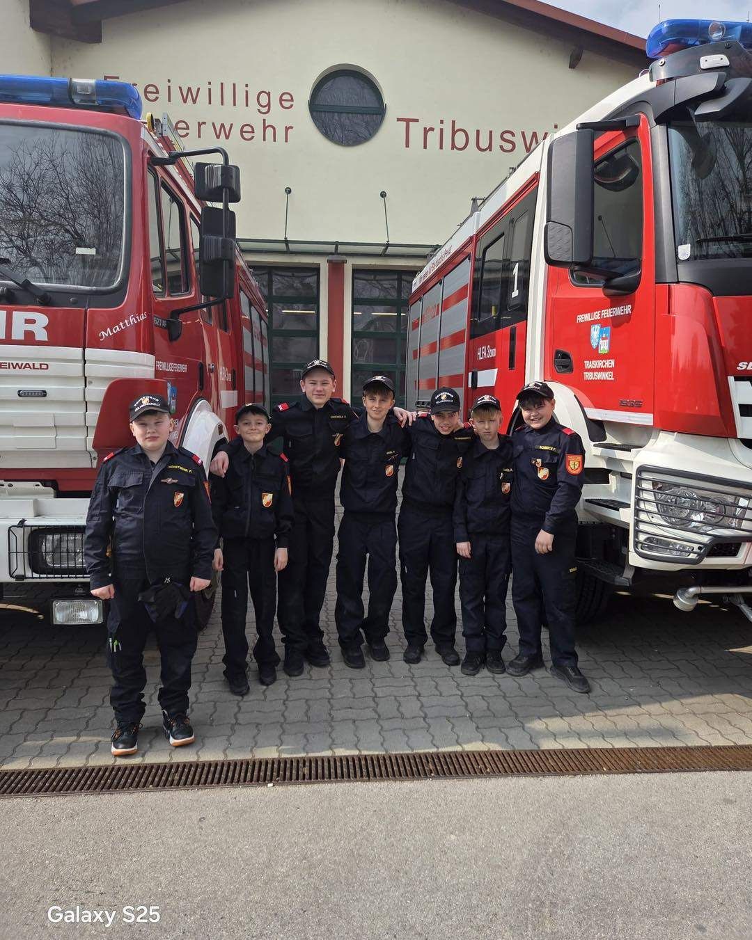 Eine Gruppe junger Leute in Uniformen steht vor zwei roten Feuerwehrwagen. Sie alle lächeln. Dahinter steht ein Gebäude mit dem Wort 'Tribuswi' darauf.