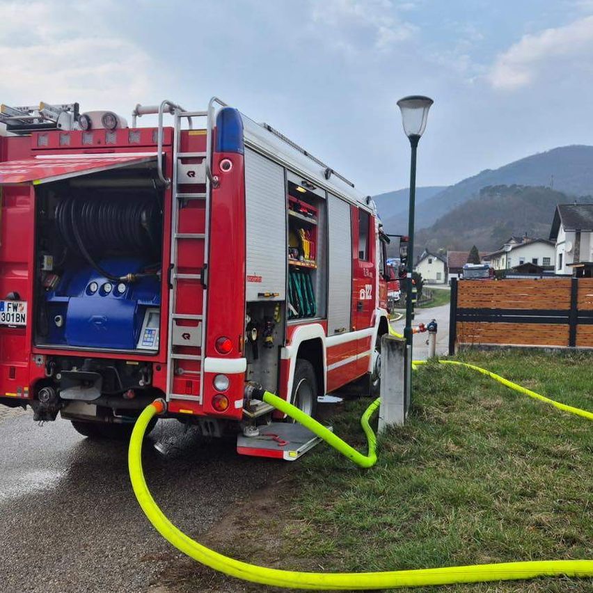 Ein roter Feuerwehrwagen ist mit offenem Heck geparkt und über gelbe Schläuche mit einer Wasserquelle verbunden auf einem Rasen.