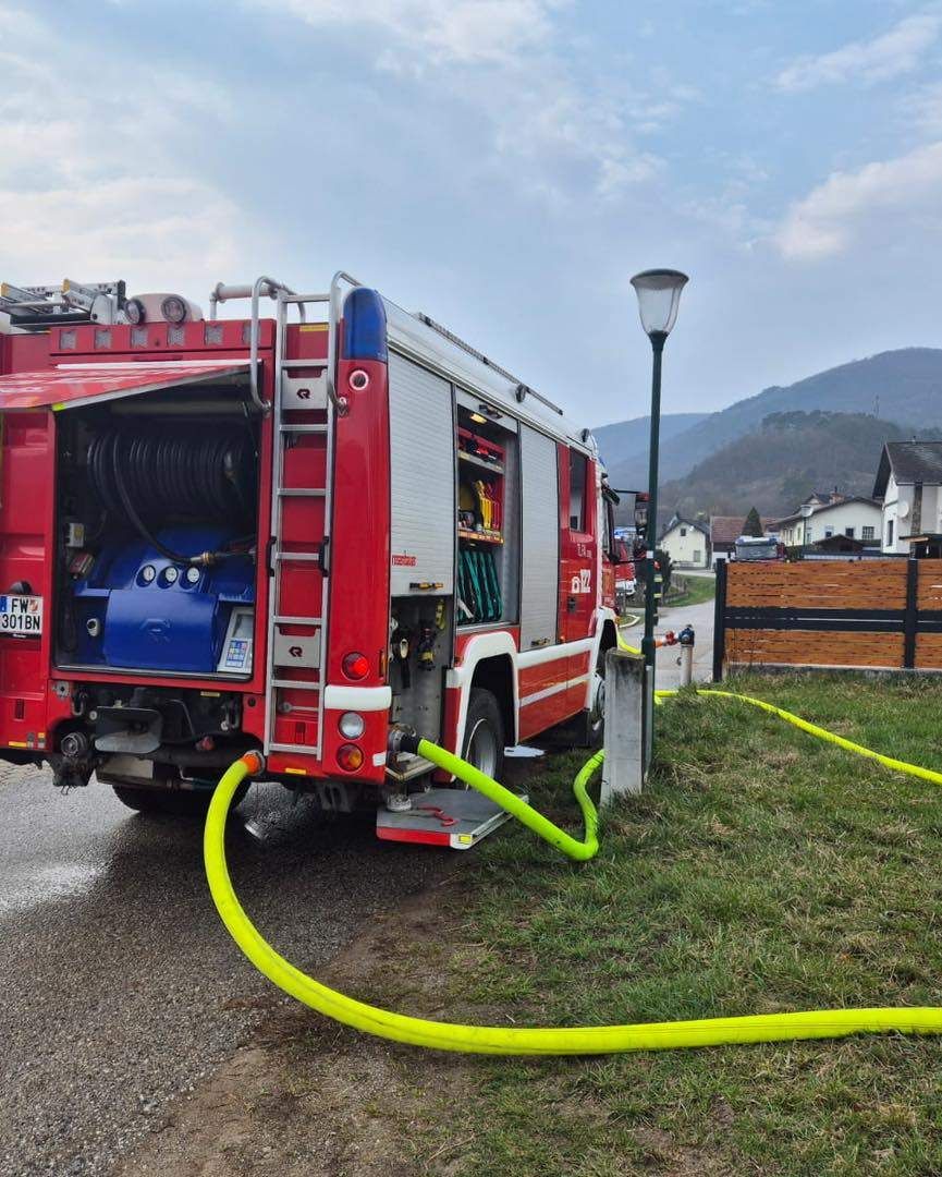 Ein roter Feuerwehrwagen ist mit offenem Heck geparkt und über gelbe Schläuche mit einer Wasserquelle verbunden auf einem Rasen.
