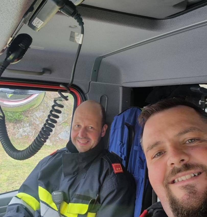 Zwei Männer sitzen in einem Fahrzeug, wahrscheinlich einem Feuerwehrauto. Einer trägt die Uniform eines Feuerwehrmanns, und beide lächeln für die Kamera.