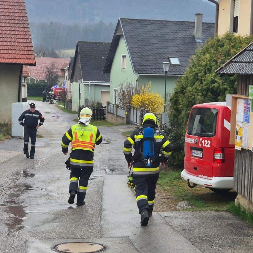 Zwei Feuerwehrleute in gelben und schwarzen Uniformen laufen auf einem nassen Bürgersteig. Ein Mann in schwarzer Uniform steht hinter ihnen. Ein roter Van ist auf der Seite geparkt.