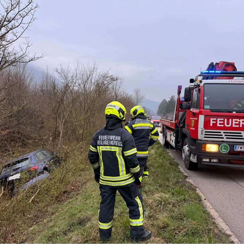Zwei Feuerwehrleute mit gelben Helmen stehen neben einem demoliert Auto am Straßenrand. Ein roter Feuerwehrwagen ist in der Nähe geparkt.