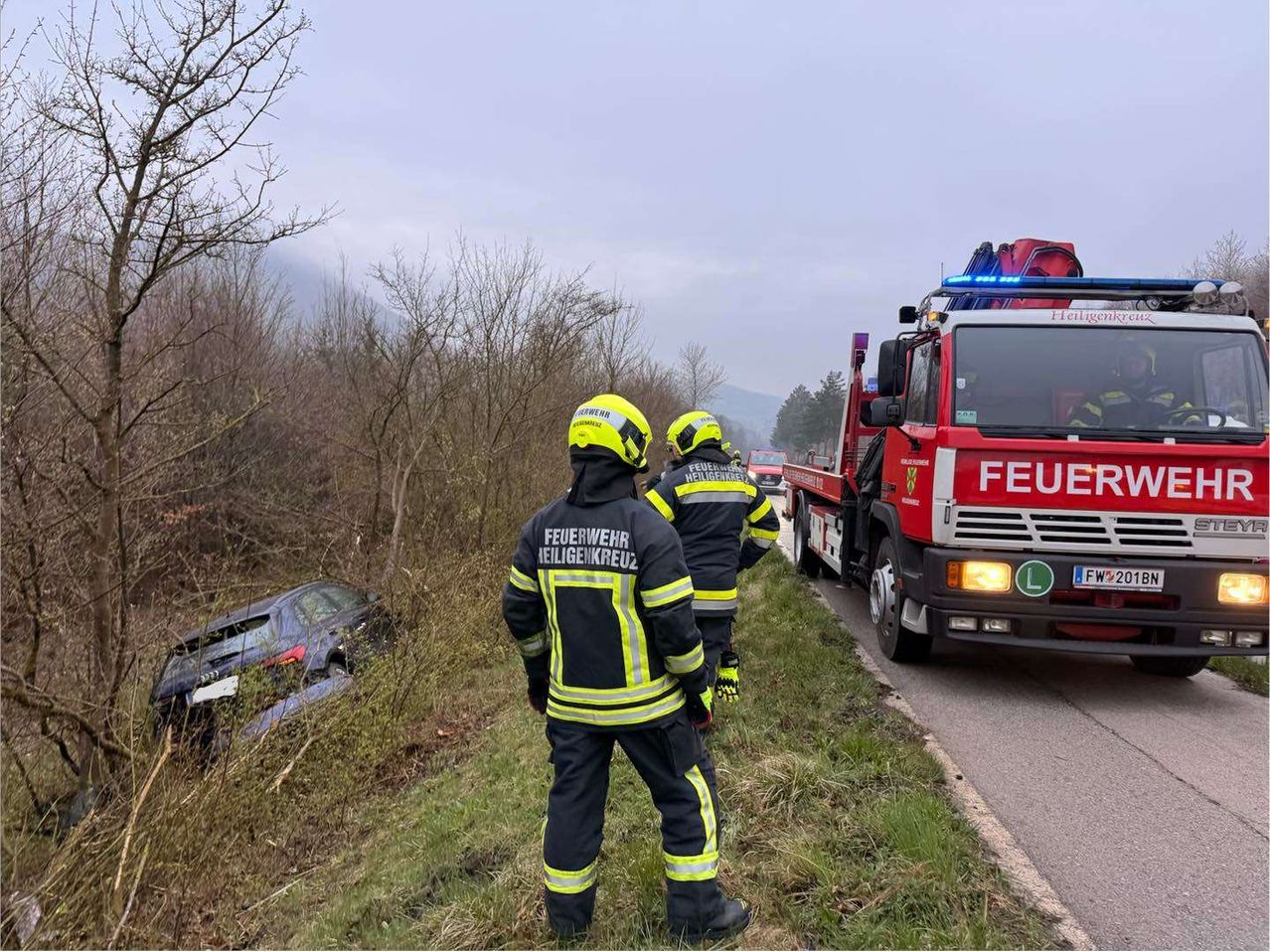 Zwei Feuerwehrleute mit gelben Helmen stehen neben einem demoliert Auto am Straßenrand. Ein roter Feuerwehrwagen ist in der Nähe geparkt.