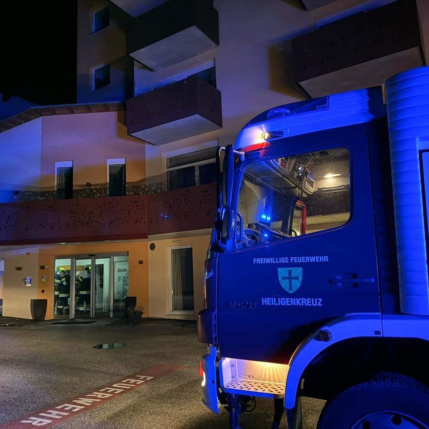 Ein blauer Feuerwehrwagen mit Heiligenkreuz-Markierungen ist vor einem Gebäude geparkt, wobei Einsatzkräfte eintreten.