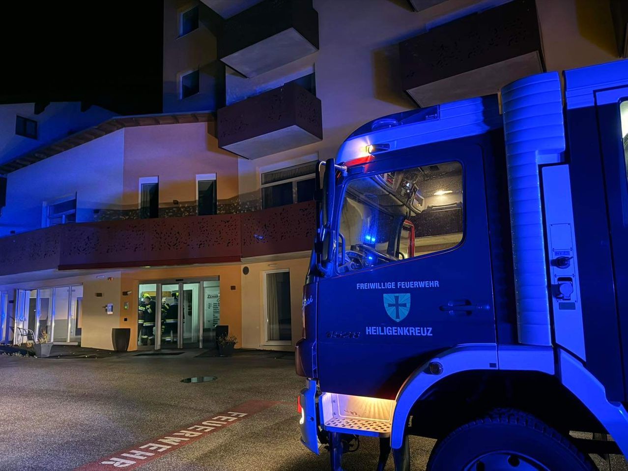 Ein blauer Feuerwehrwagen mit Heiligenkreuz-Markierungen ist vor einem Gebäude geparkt, wobei Einsatzkräfte eintreten.