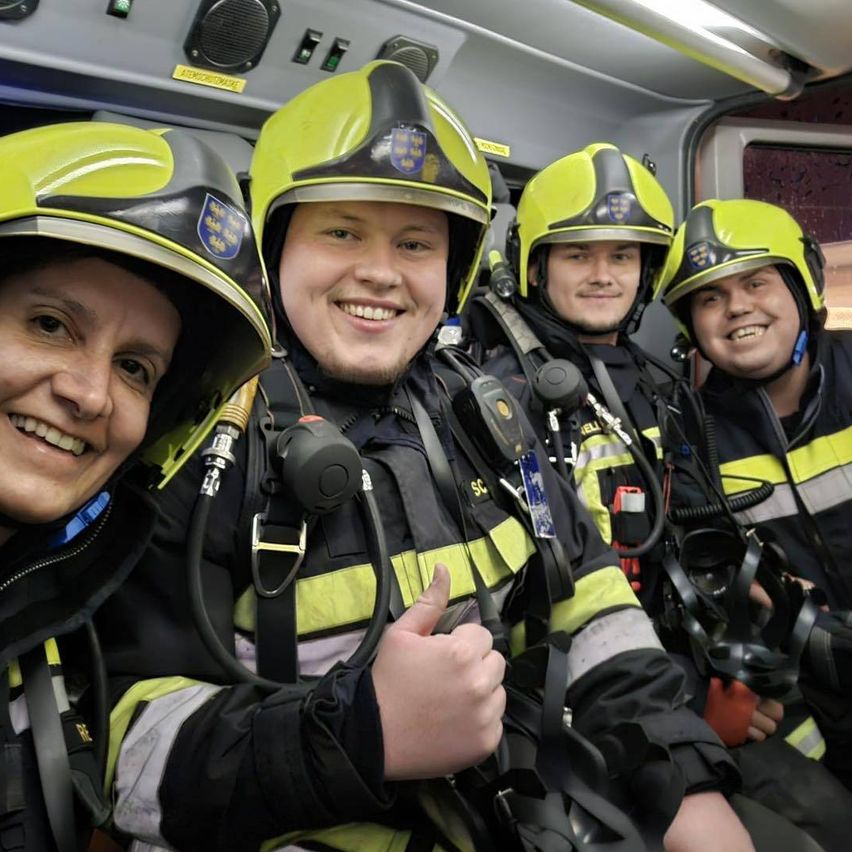 Fünf Feuerwehrleute in voller Ausrüstung, lächelnd, sitzen in einem Fahrzeug. Die Helme haben ein Logo und gelbe Streifen. Eine Person gibt einen Daumen hoch.