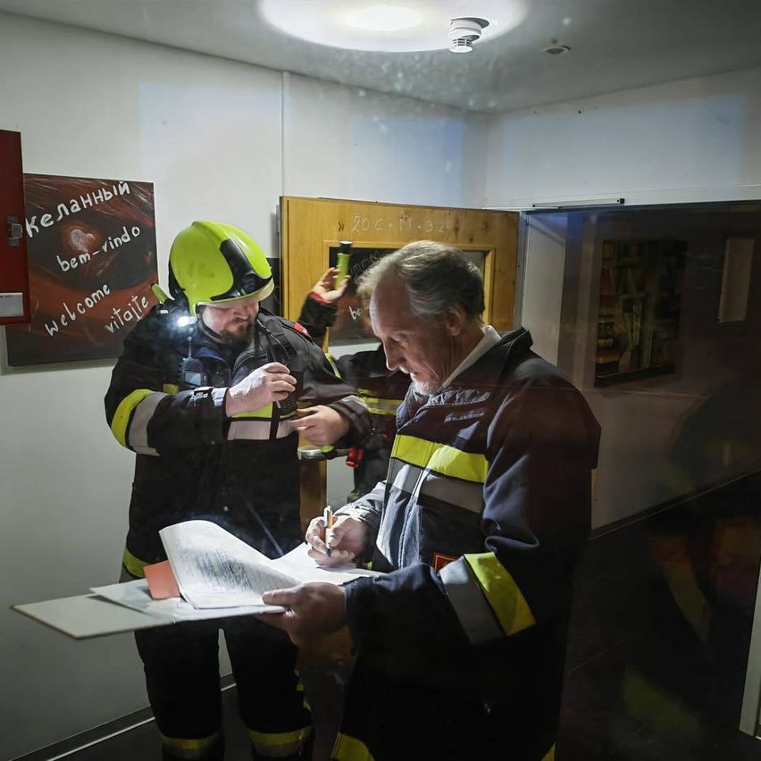 Zwei Feuerwehrleute überprüfen Papierkram in einem Raum mit einer Holztür. Einer hält einen Stift und Papier, während der andere mit einer Taschenlampe steht. Ein Schild an der Wand sagt 'Willkommen', und ein Regal ist im Hintergrund sichtbar.