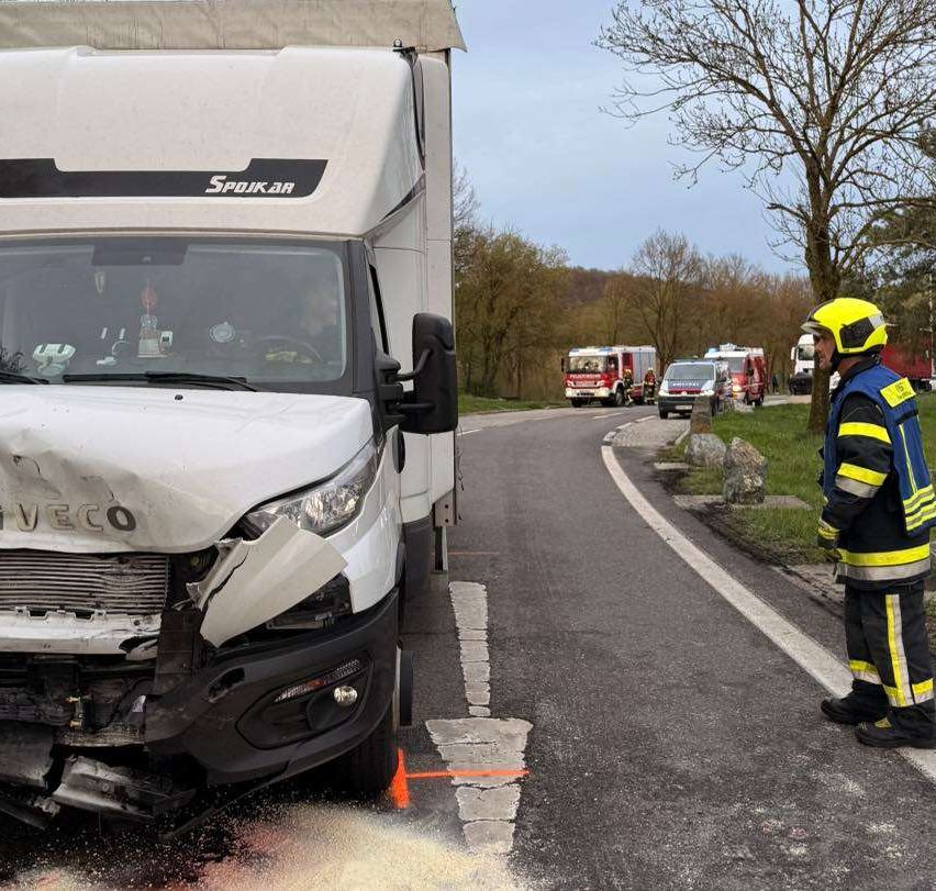 Ein weißer LKW mit beschädigter Front steht am Straßenrand. Ein Feuerwehrmann steht in der Nähe des LKW. Andere Rettungsfahrzeuge sind in der Nähe geparkt.