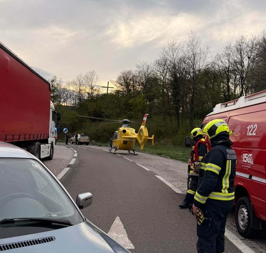 Ein gelber Hubschrauber ist am Straßenrand gelandet. Ein rotes Notfallfahrzeug und zwei Feuerwehrleute stehen in der Nähe. Ein Lkw und ein Auto sind ebenfalls vorhanden. Bäume und ein Kreuz sind im Hintergrund zu sehen.