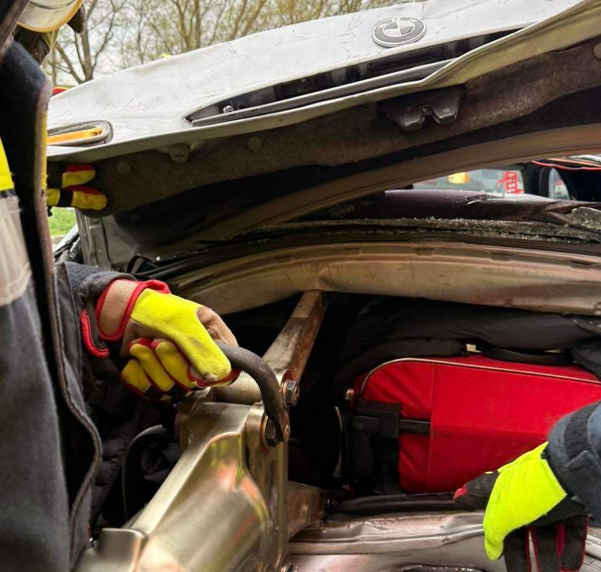 Eine Person in Schutzkleidung arbeitet am Motor eines beschädigten Autos. Die Motorhaube ist offen und zeigt den Motor. Eine rote Tasche befindet sich im Inneren des Autos, neben einem schwarzen Gegenstand.