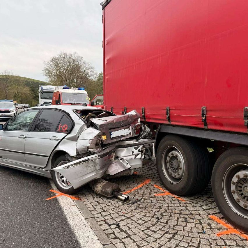 Ein silbernes Auto ist nach einer Kollision mit einem roten Lastwagen auf einer asphaltierten Straße schwer beschädigt. Rettungsfahrzeuge sind vor Ort.