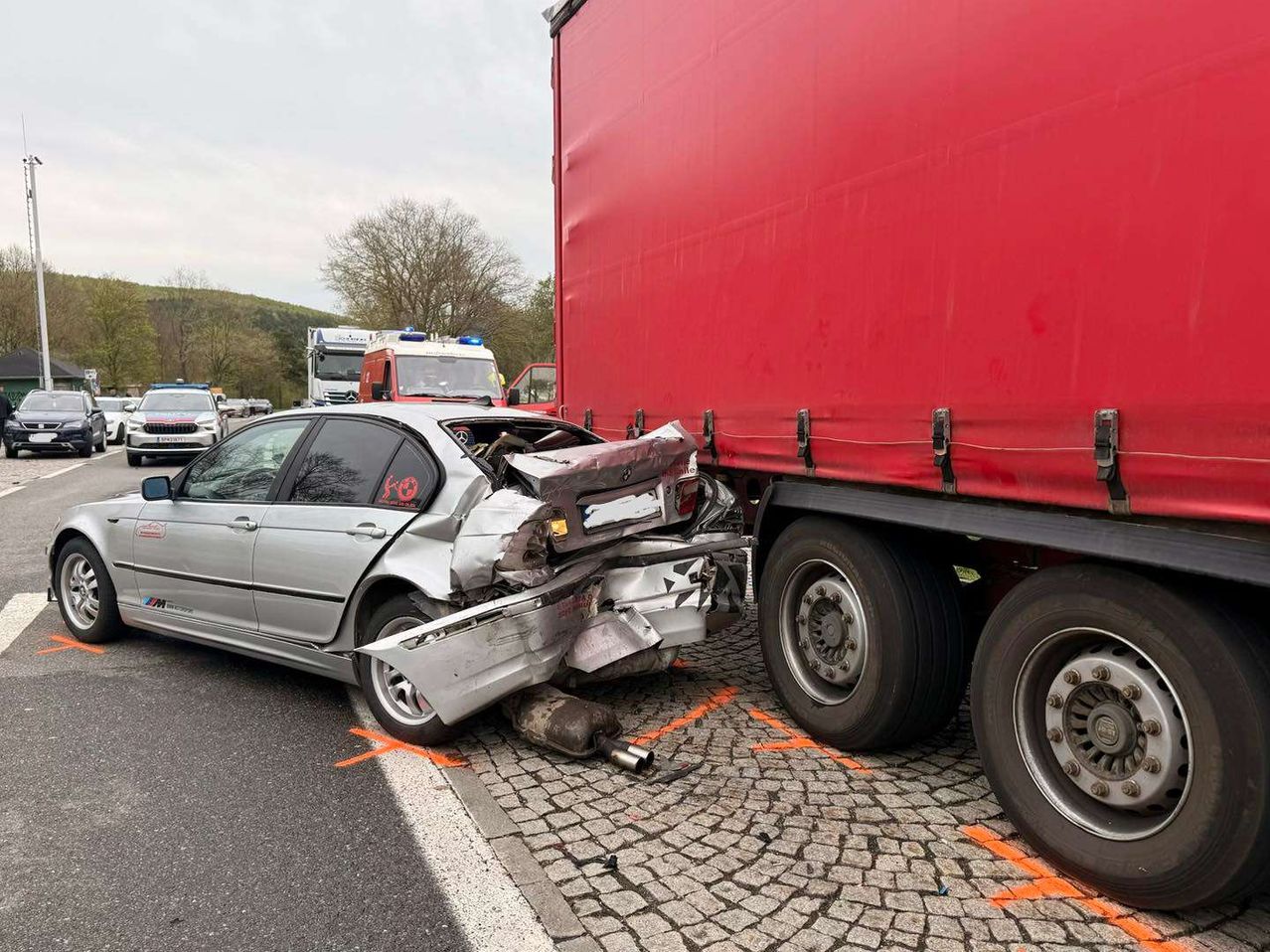 Ein silbernes Auto ist nach einer Kollision mit einem roten Lastwagen auf einer asphaltierten Straße schwer beschädigt. Rettungsfahrzeuge sind vor Ort.