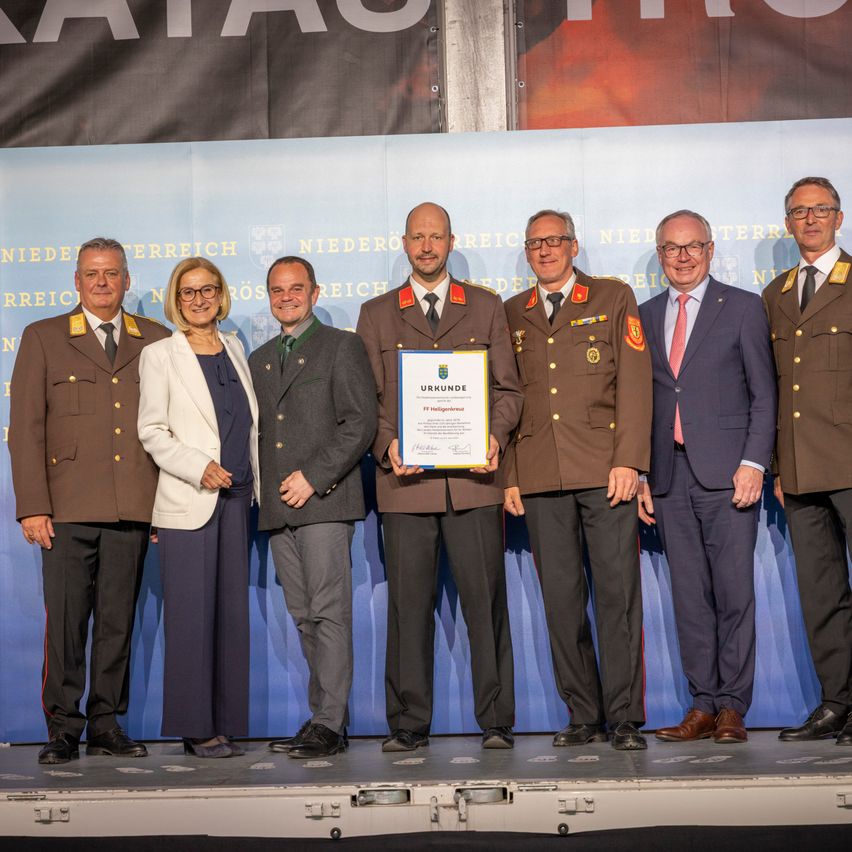 Mehrere Personen in Uniform, darunter zwei Frauen, stehen auf der Bühne und halten ein Zertifikat. Hinter ihnen befindet sich ein blauer Hintergrund mit Text.