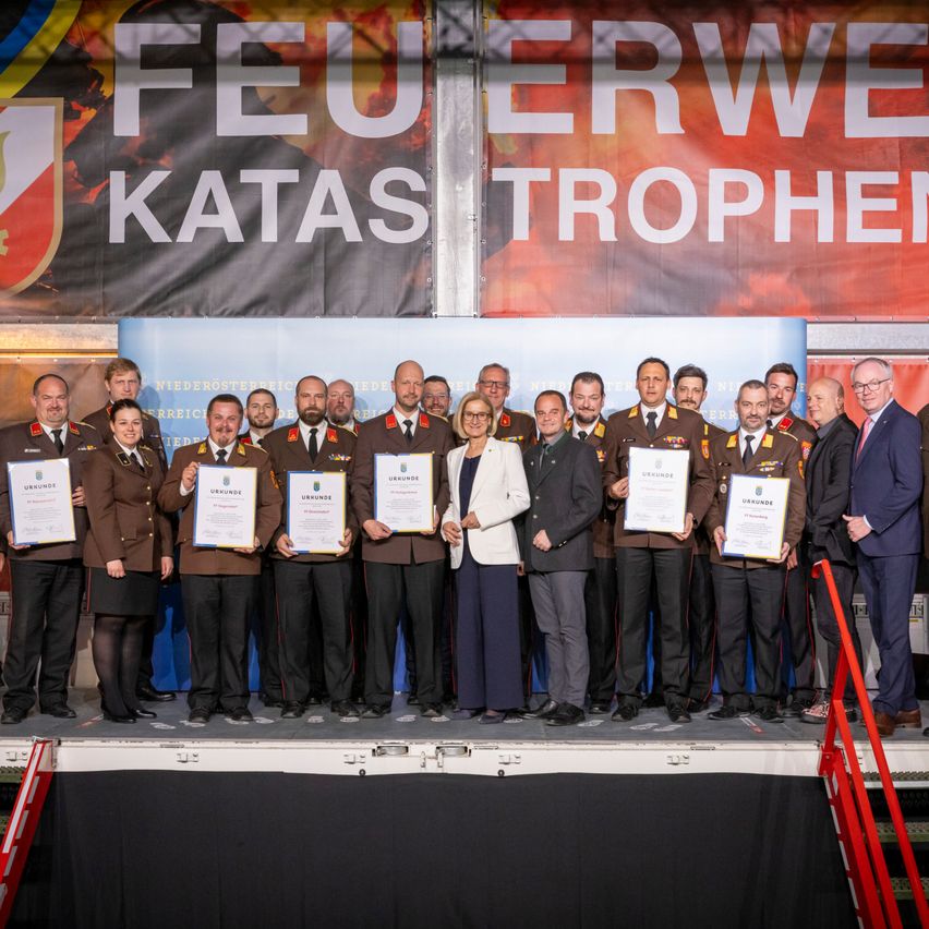 Eine Gruppe von Menschen in Uniform, darunter Militär und Zivilisten, stehen auf einer Bühne und halten Urkunden in der Hand. Ein Banner hinter ihnen lautet 'Feuerwehr Katas Trophen'.