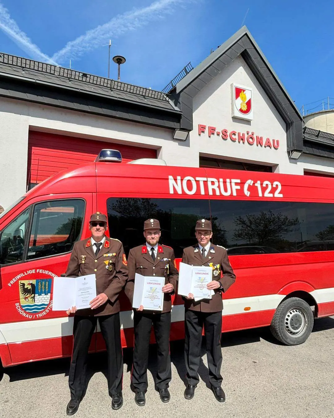 Drei Männer in Uniform stehen vor einem roten Fahrzeug mit der Aufschrift Notruf 122. Sie halten Urkunden in der Hand und lächeln. Hinter ihnen befindet sich ein Gebäude mit dem Logo FF-Schönau.
