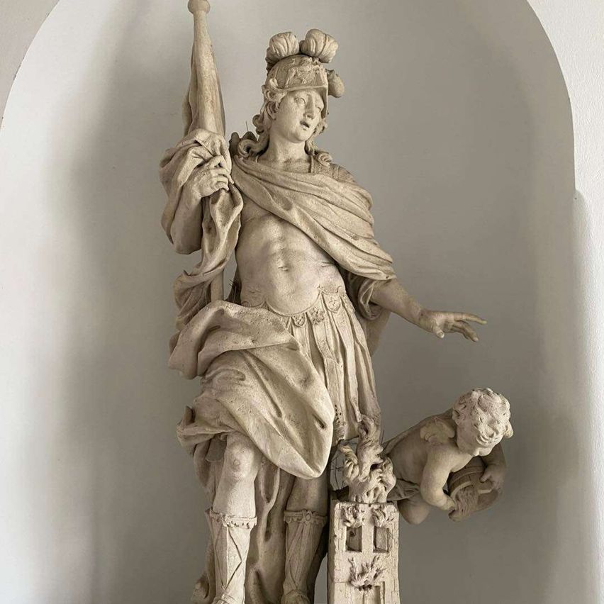 Eine Marmorstatue einer männlichen Figur mit Lorbeerkranz, die eine Fahne hält, mit einem kleinen Cherub darunter.