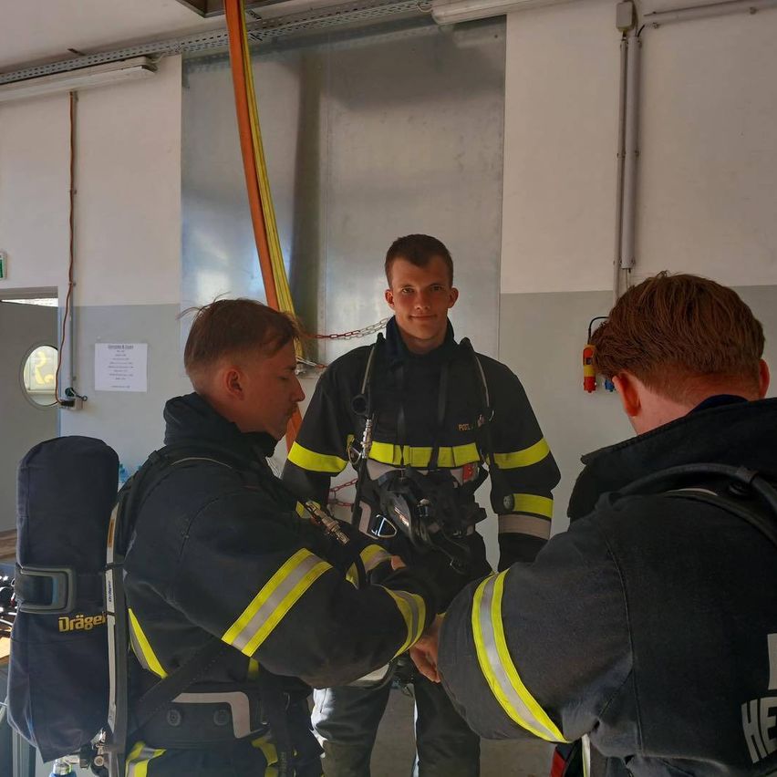 Drei Feuerwehrleute in voller Montur stehen in einem Raum. Einer hält eine Maske, ein anderer greift nach seiner Hand und der dritte schaut ihn an.