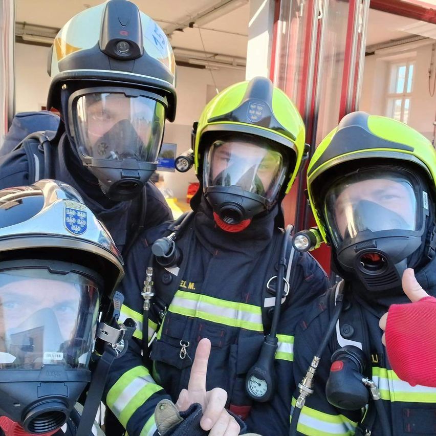 Eine Gruppe von Feuerwehrleuten in Schutzausrüstung mit Helmen steht in einer Feuerwache. Sie tragen Masken und posieren für ein Foto.