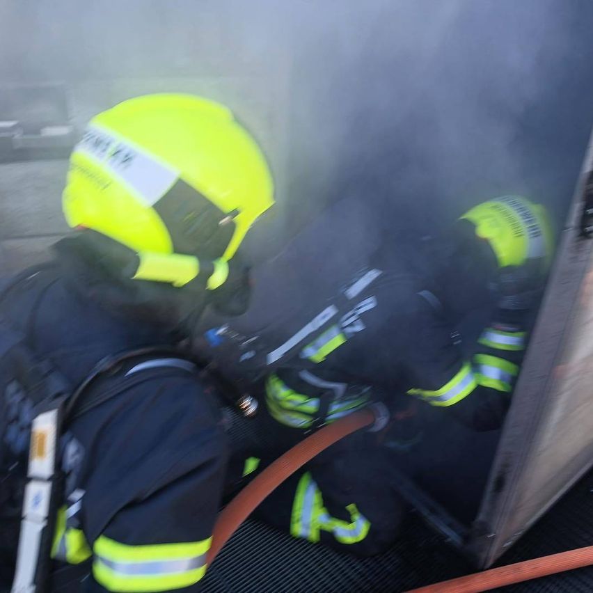 Feuerwehrleute in Schutzausrüstung löschen einen Brand mit einem Schlauch. Ein Feuerwehrmann steht am Boden, der andere steht. Rauch füllt die Luft.