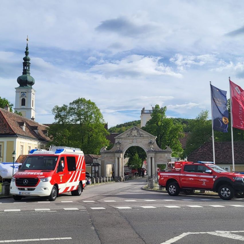Ein rot-weißer Feuerwehrwagen und ein roter Pickup-Truck stehen vor einem historischen Gebäude mit Flaggen an Masten.