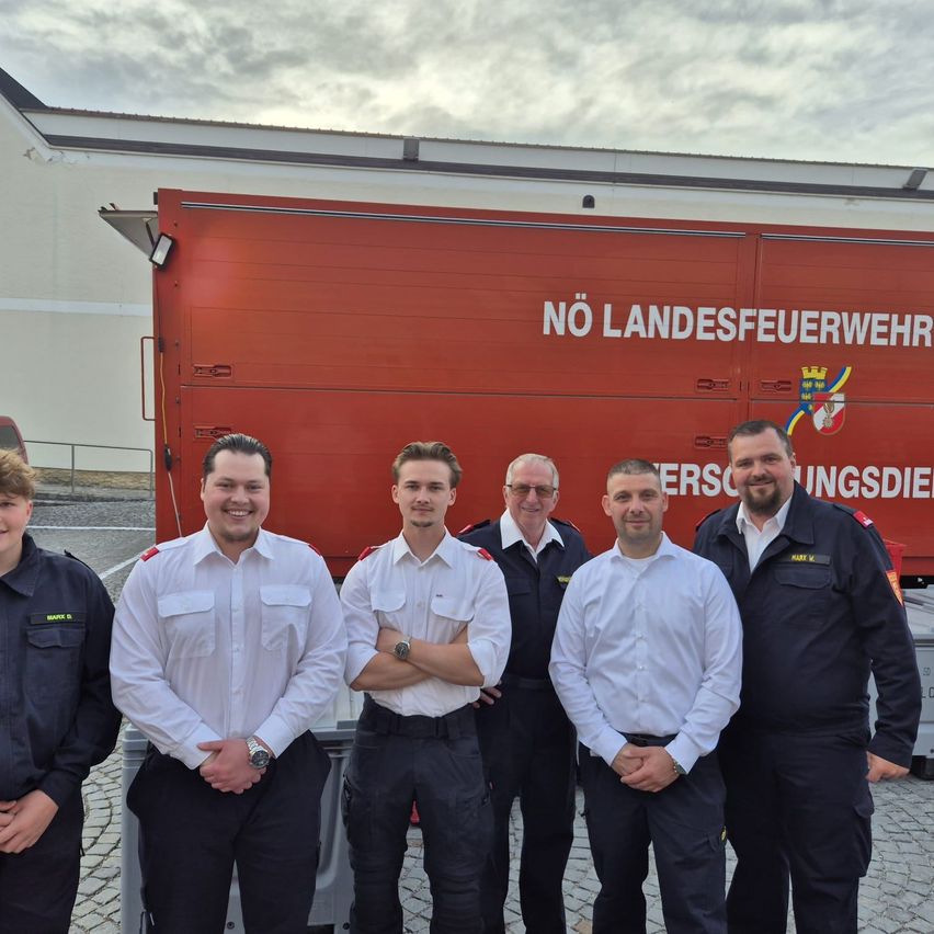 Fünf Feuerwehrleute posieren vor einem roten Lastwagen mit der Aufschrift 'NÖ LANDESFEUERWEHR'. Sie tragen Uniformen und lächeln für das Foto. Hinter ihnen steht ein Gebäude mit einem bewölkten Himmel.