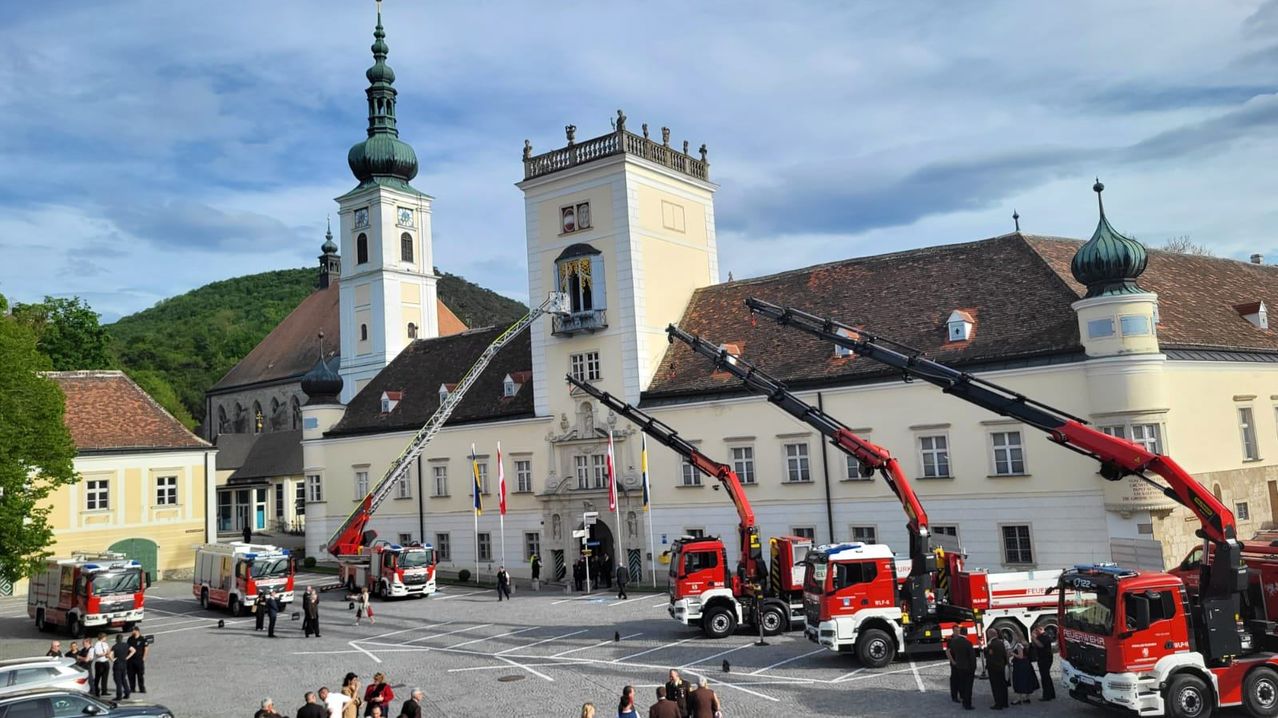 Ein Feuerwehrfahrzeug mit ausgefahrener Leiter steht vor einem historischen Gebäude mit mehreren Fahnen, und zwei weitere Feuerwehrfahrzeuge sind in der Nähe geparkt. Menschen haben sich in der Gegend versammelt.