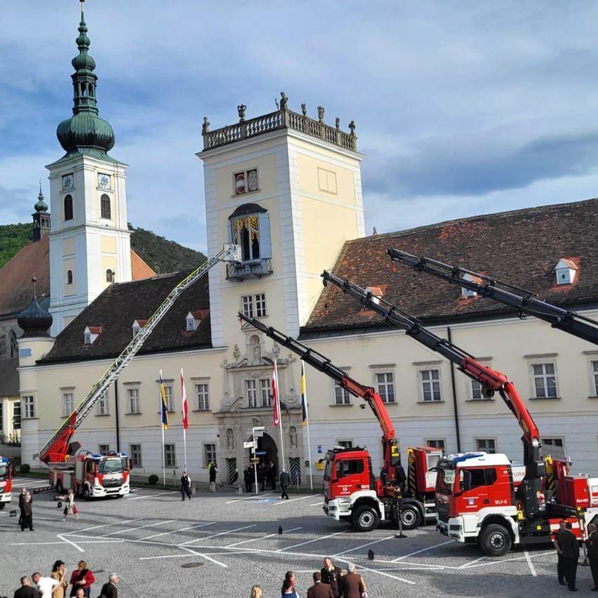 Ein Feuerwehrfahrzeug mit ausgefahrener Leiter steht vor einem historischen Gebäude mit mehreren Fahnen, und zwei weitere Feuerwehrfahrzeuge sind in der Nähe geparkt. Menschen haben sich in der Gegend versammelt.