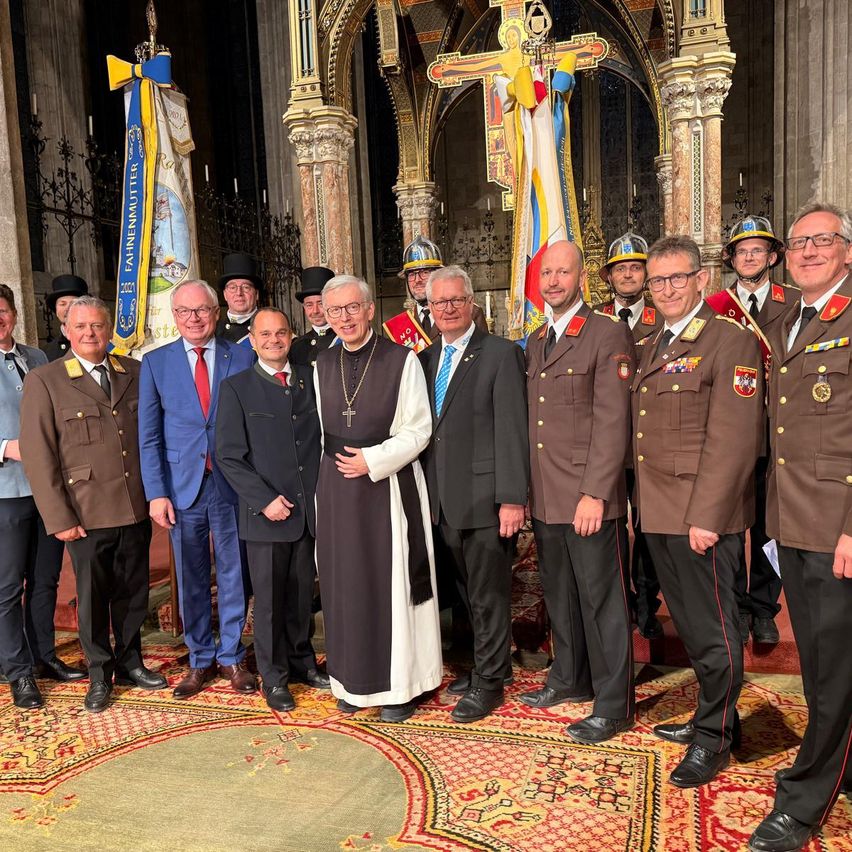 Eine Gruppe von Männern in formeller Kleidung, darunter ein Priester, steht vor einem Kircheninneren mit einem Kreuz und Flaggen.