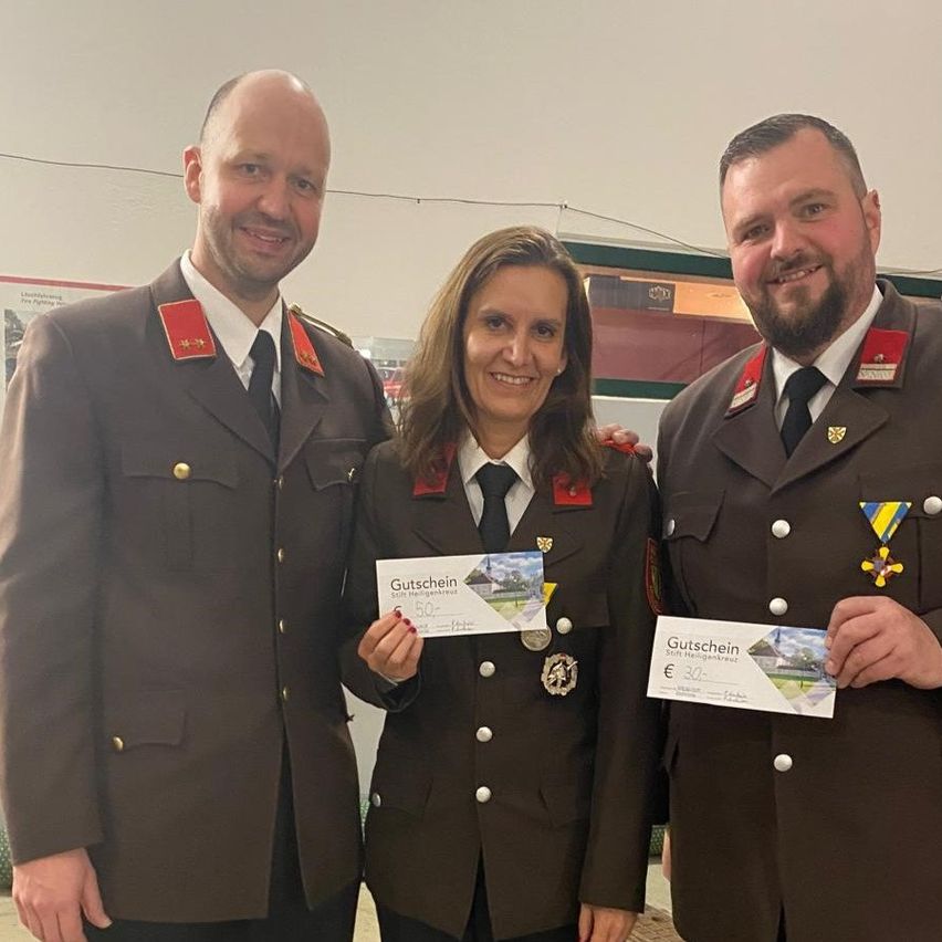 Drei Menschen in Uniform, zwei Männer und eine Frau, stehen eng beieinander und lächeln für ein Foto. Die Frau hält einen Geschenkgutschein.