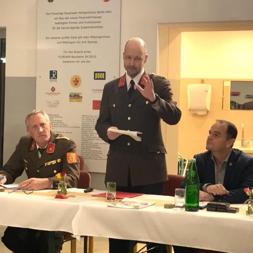 Drei Männer sitzen an einem Tisch; einer in Uniform gestikuliert, ein anderer hört aufmerksam zu, und der dritte sieht nachdenklich aus. Hinter ihnen steht ein Banner mit der Aufschrift 'Floriani-Bausteine in Gold'. Eine Glasflasche und ein Papierbecher stehen auf dem Tisch.