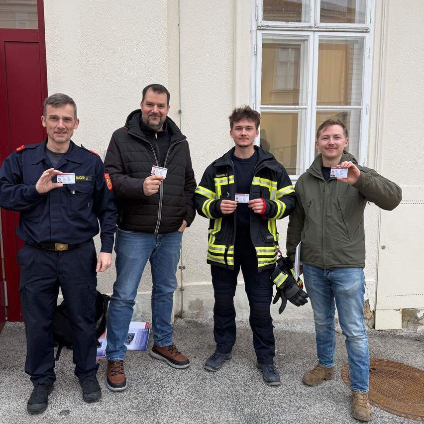 Vier Männer stehen vor einem Gebäude mit einer roten Tür und lächeln. Jeder hält eine Karte in der Hand, wahrscheinlich Ausweise. Einer trägt einen Feuerwehrmannsmantel.
