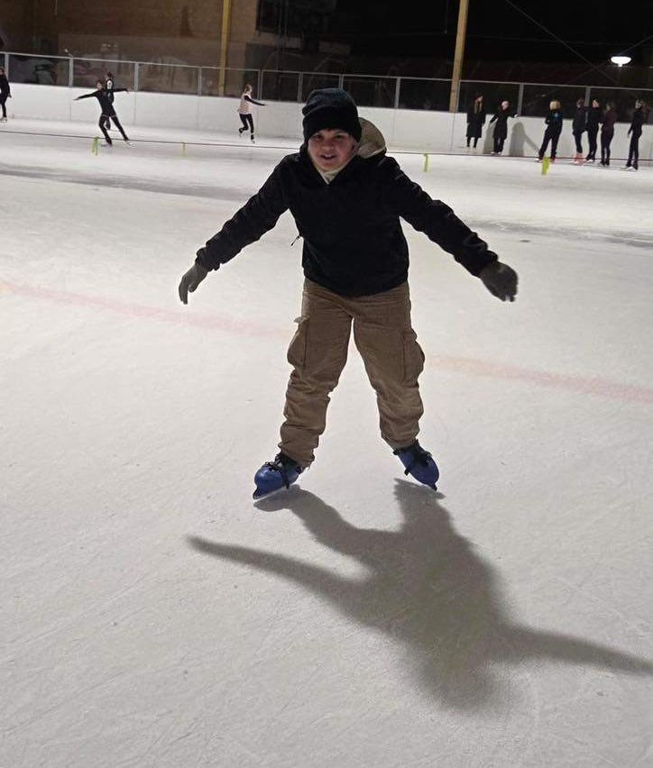 Ein Junge eiskunstlauft auf einer Eisbahn, gekleidet in einem schwarzen Jacken, braunen Hosen und Eislaufschuhen. Hinter ihm eiskunstlaufen mehrere Personen.
