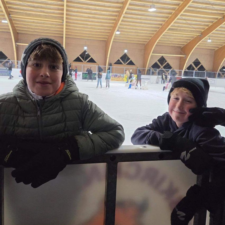 Zwei junge Jungen stehen an der Barriere einer Eishalle. Sie lächeln in die Kamera. Im Hintergrund sind mehrere Menschen beim Eislaufen zu sehen.