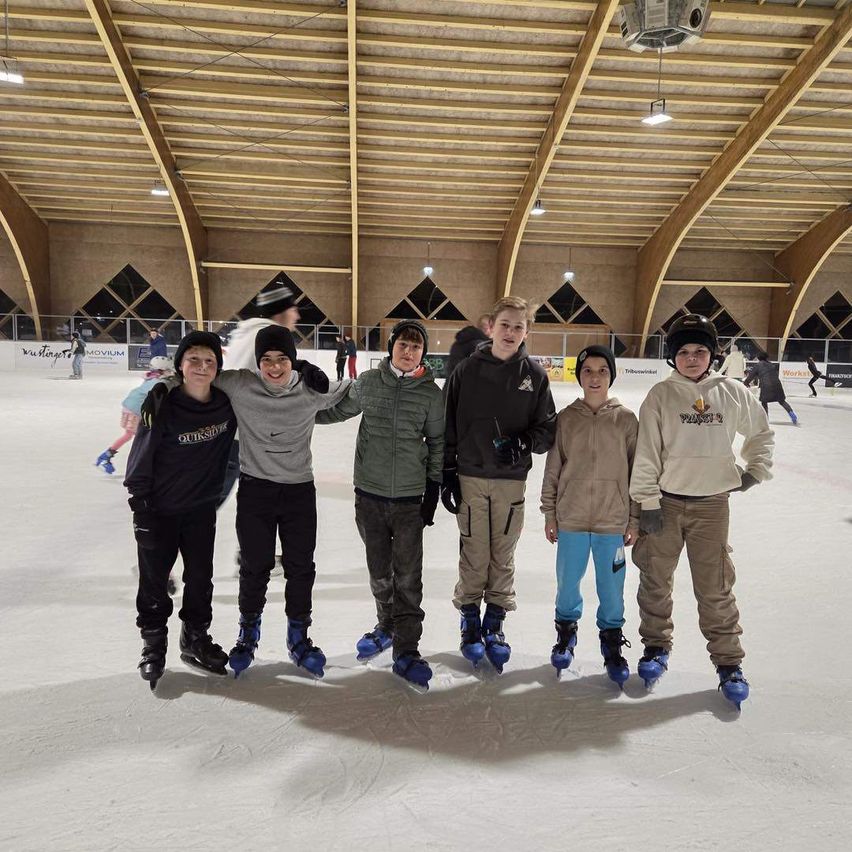 Eine Gruppe von fünf Jungen in Winterkleidung steht in einer überdachten Eishalle, lächelt und posiert für ein Foto.
