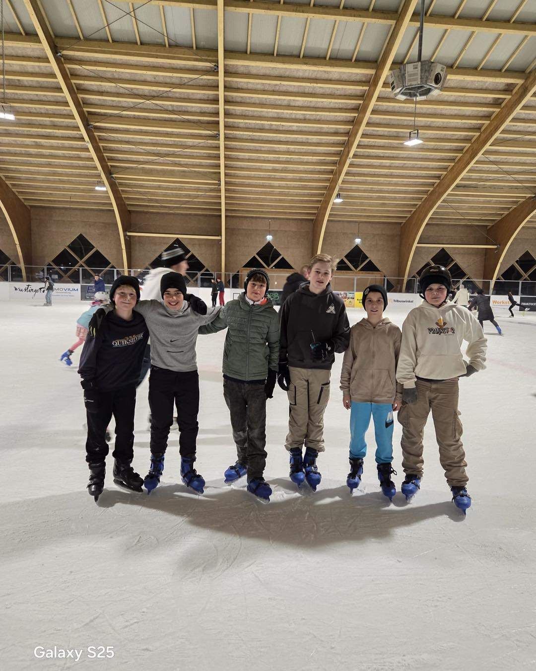 Eine Gruppe von fünf Jungen in Winterkleidung steht in einer überdachten Eishalle, lächelt und posiert für ein Foto.