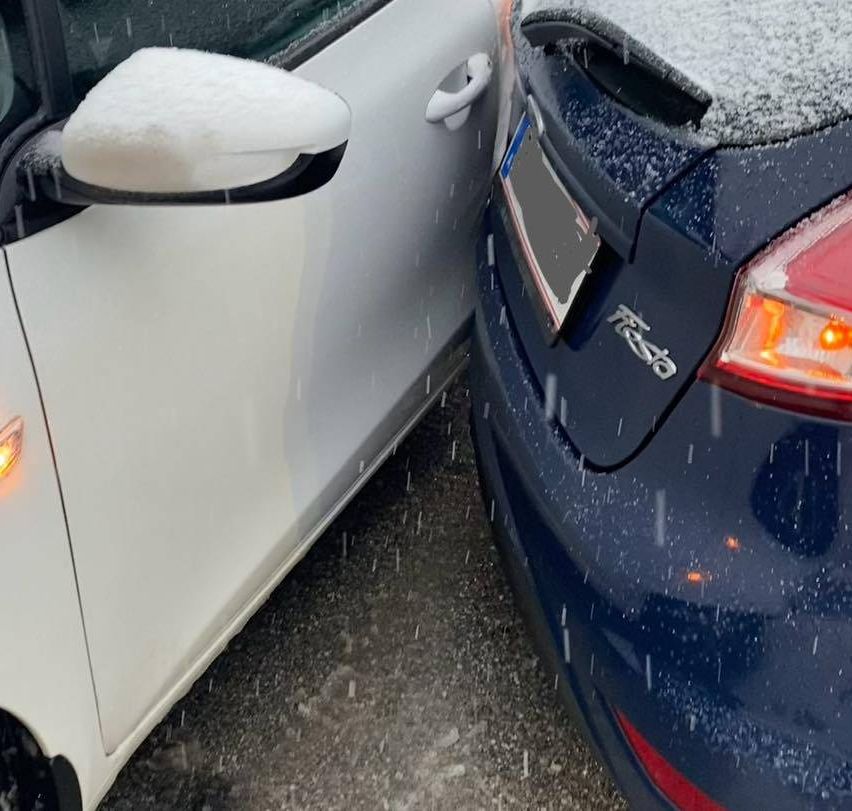Zwei Autos sind geparkt, eines weiß und eines blau. Das weiße Auto hat Schnee auf seinem Spiegel, und das blaue Auto hat Eis auf seinem Heck.