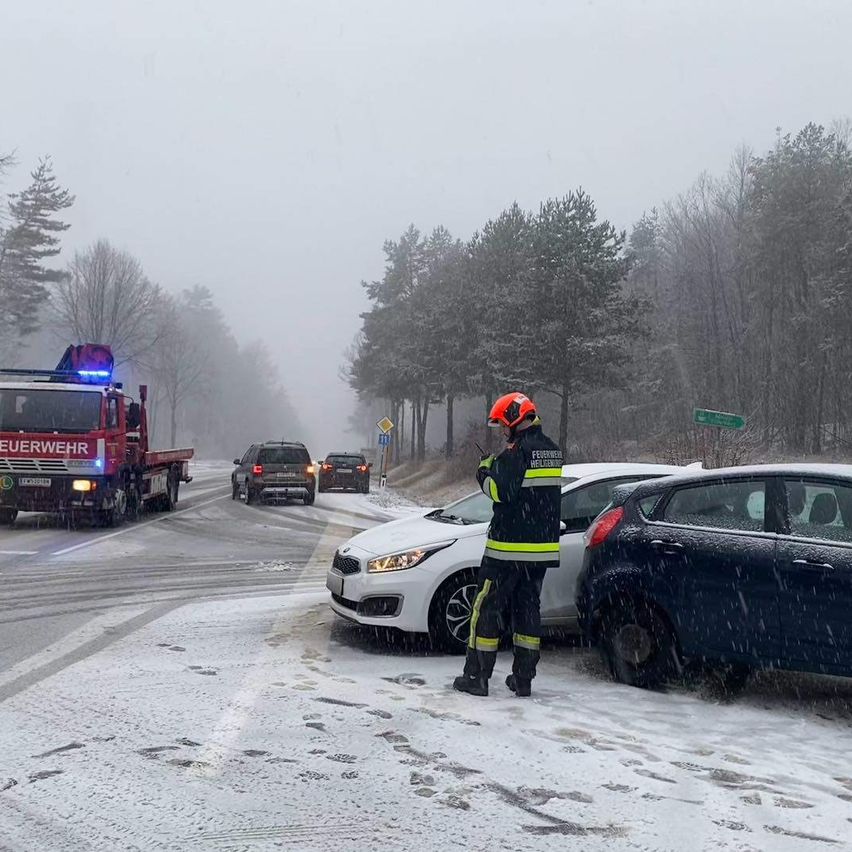Rettungskräfte auf einer verschneiten Straße. Ein Feuerwehrmann steht neben einem beschädigten Auto. Ein Feuerwehrwagen und mehrere Autos sind im Hintergrund.