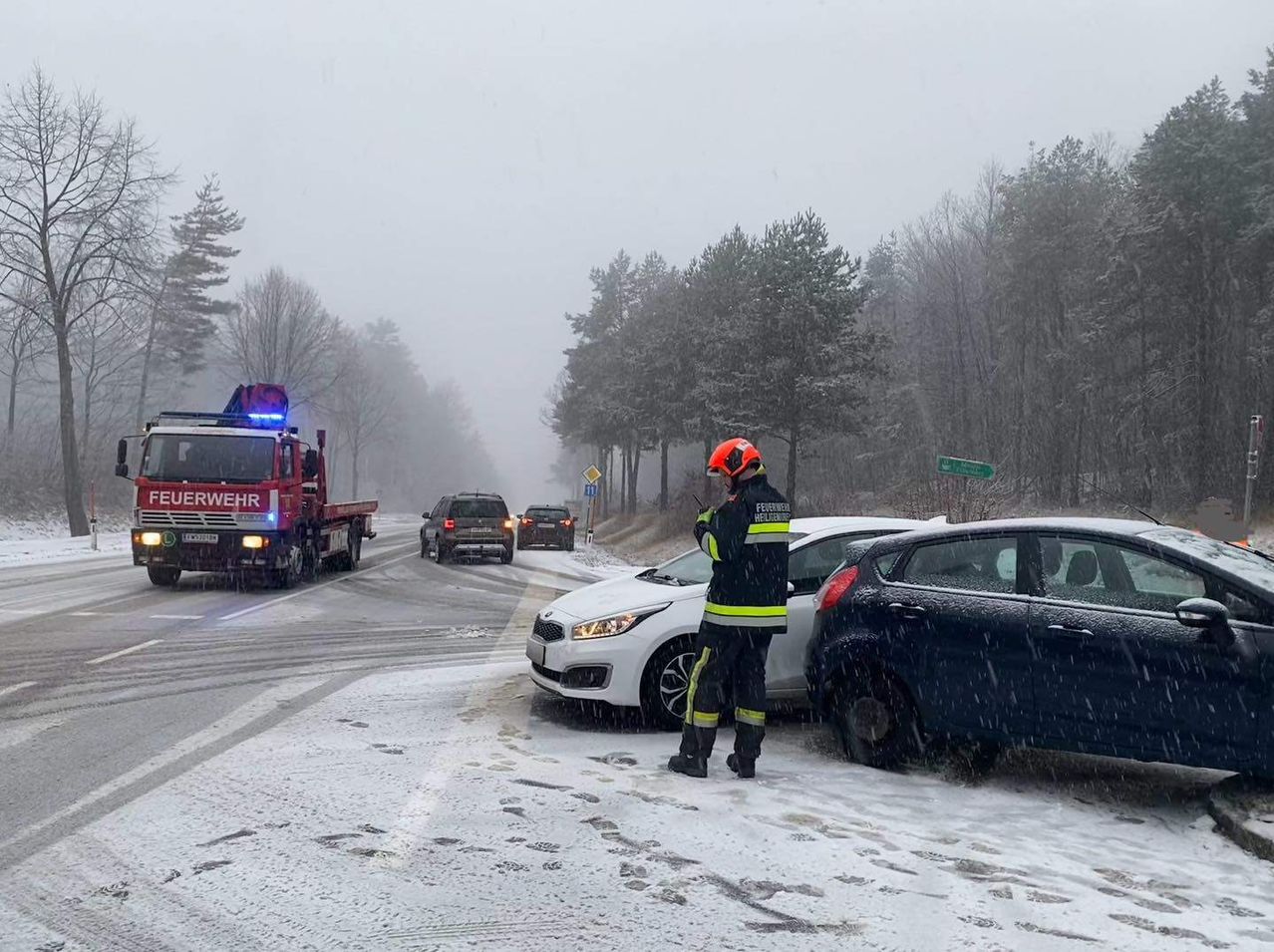Rettungskräfte auf einer verschneiten Straße. Ein Feuerwehrmann steht neben einem beschädigten Auto. Ein Feuerwehrwagen und mehrere Autos sind im Hintergrund.