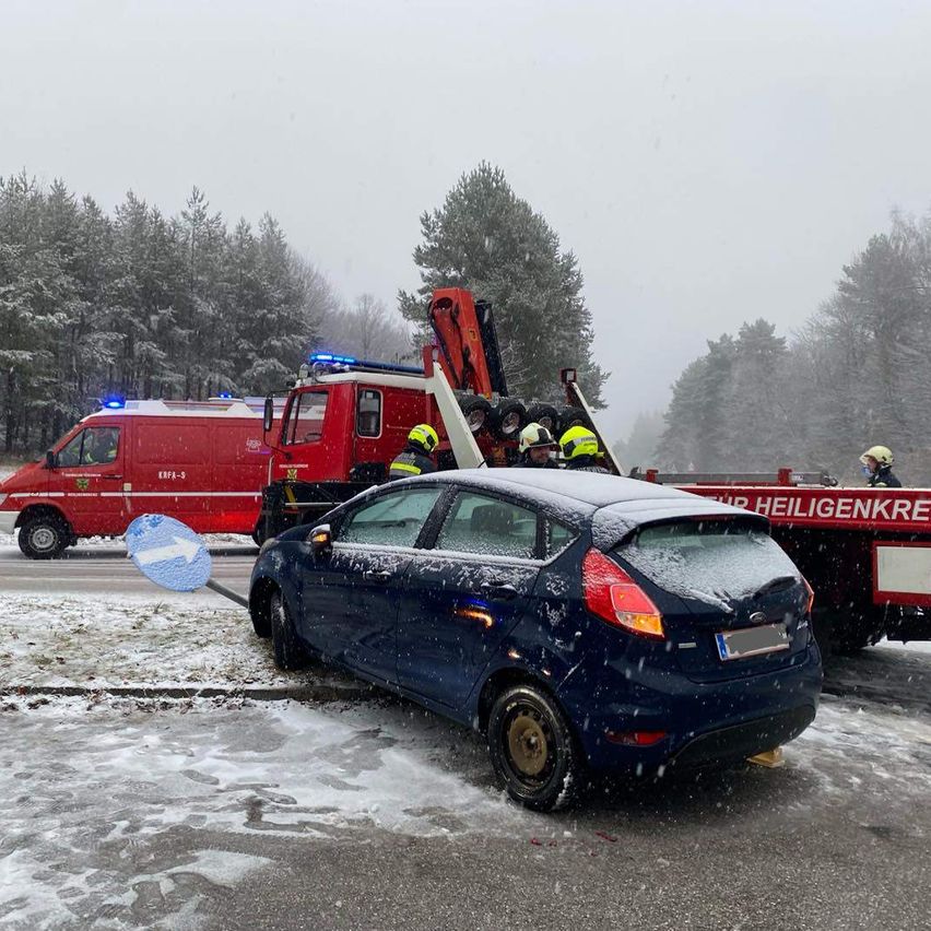 Ein blaues Auto mit einem beschädigten Stoßfänger wird von einem roten Abschleppwagen weggezogen. Zwei Personen in gelben Helmen arbeiten am Auto. Schnee liegt auf dem Boden und an den Bäumen.