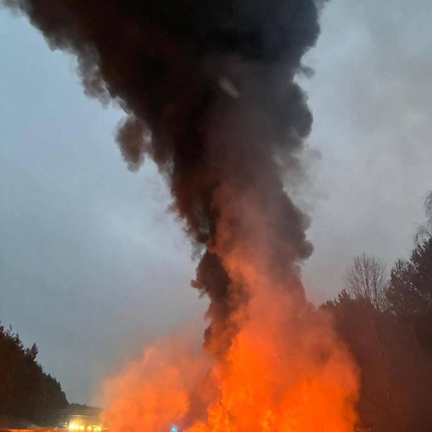 Ein großes Feuer brennt auf der Straße und produziert dicken schwarzen Rauch. Die Straße ist von Bäumen umgeben, und es gibt ein Gebäude auf der linken Seite.