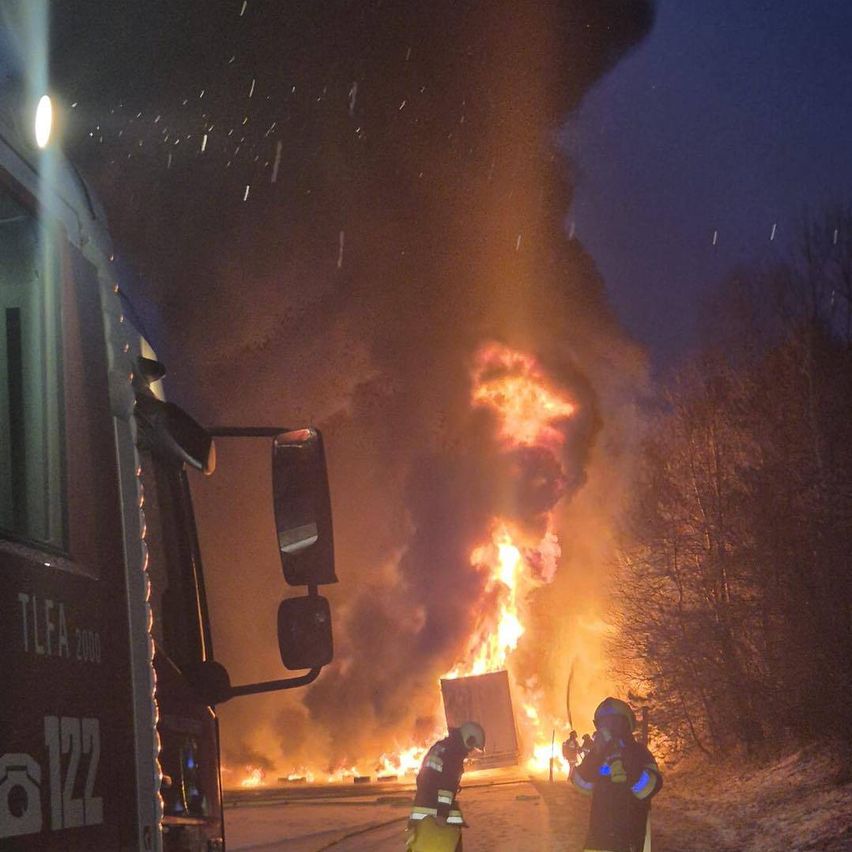 Ein feuerspeiender LKW steht bei Nacht im Brand. Zwei Feuerwehrleute in voller Ausrüstung stehen neben dem Fahrzeug.