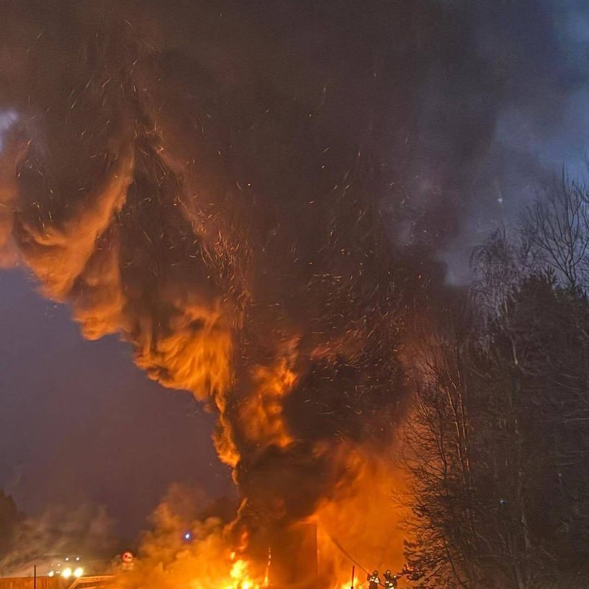 Ein großes Feuer umgibt nachts ein Gebäude, während Feuerwehrleute versuchen, die Flammen zu löschen. Rauch und Funken steigen aus dem Feuer auf.
