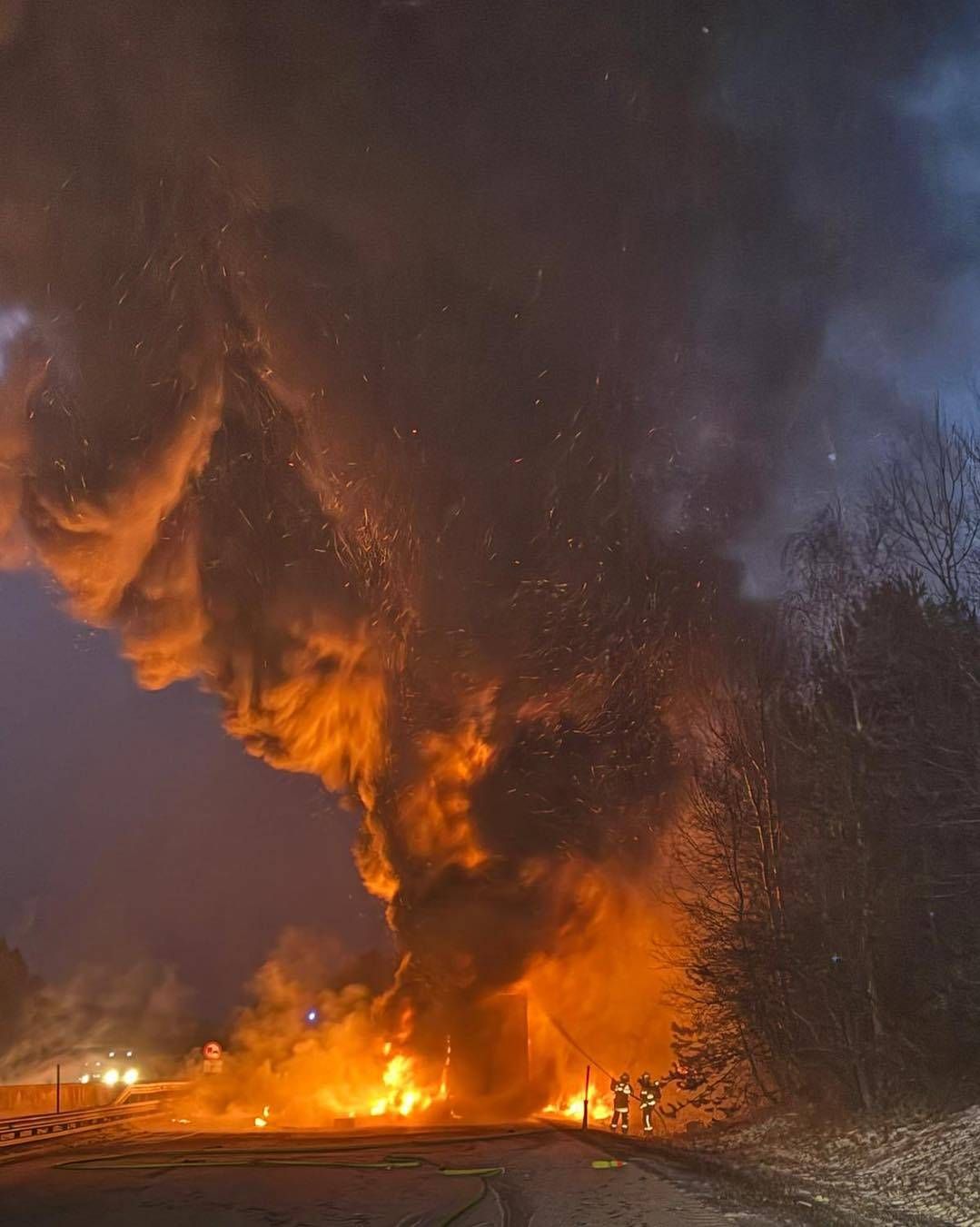 Ein großes Feuer umgibt nachts ein Gebäude, während Feuerwehrleute versuchen, die Flammen zu löschen. Rauch und Funken steigen aus dem Feuer auf.