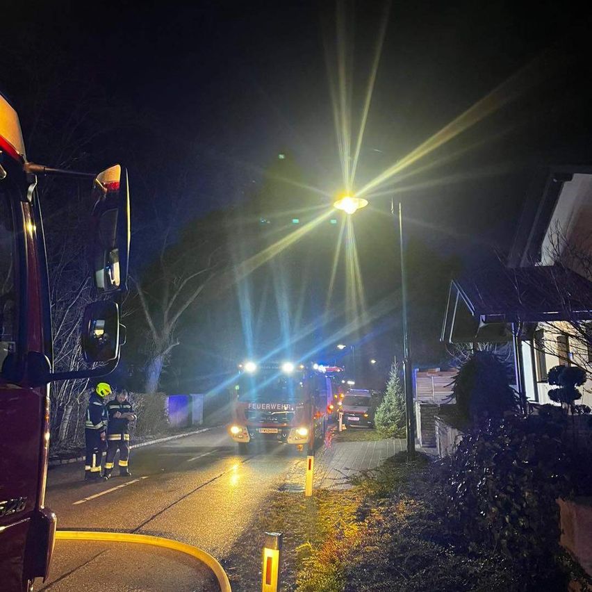 Zwei Feuerwehrleute stehen neben einem Feuerwehrauto auf einer dunklen Straße bei Nacht, mit einem Haus und Straßenlaternen im Hintergrund.