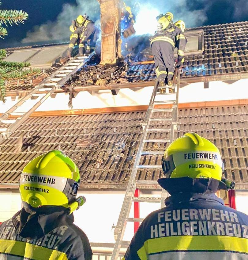 Feuerwehrleute sind auf dem Dach eines Hauses mit aufsteigendem Rauch. Sie benutzen Leitern, um auf das Dach zu gelangen. Sie tragen gelbe Helme mit den Worten 'Feuerwehr' und 'Heilgenkreuz' darauf.